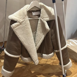 Beige teddyjacka från ASOS DESIGN - Säljer en beige teddyjacka från ASOS DESIGN med vit fuskpäls på krage, ärmslut och nederkant. Jackan är croppad och har dragkedja samt breda slag. Perfekt för kyliga dagar!