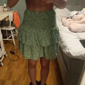 Grön blommig volangkjol - Säljer en söt grön kjol med småblommigt mönster och volanger. Kjolen har resår i midjan och är kort i modellen, perfekt för varma dagar. Lätt och luftig känsla. Går även att använda som topp