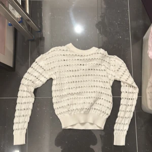 Vit stickad tröja med hålmönster - Säljer en vit stickad tröja med rund hals och dekorativt hålmönster över hela tröjan. Tröjan har långa ärmar och ribbade muddar vid ärmslut och nederkant. Perfekt för lager-på-lager-stil!