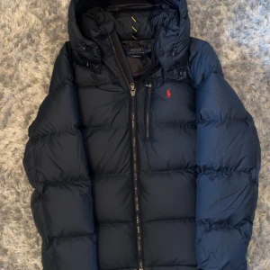 Mörkblå dunjacka från Polo Ralph Lauren - Säljer en mörkblå dunjacka från Polo Ralph Lauren med huva och dragkedja. Jackan har en röd broderad logga på bröstet och flera praktiska fickor. Perfekt för kyliga dagar och riktigt snygg streetstil.