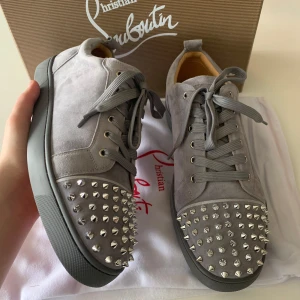 Grå mockasneakers med nitar från Christian Louboutin - Säljer ett par grå sneakers i mocka från Christian Louboutin med silverfärgade nitar framtill och klassisk röd sula. Skorna har snörning och rund tå, samt en platt sula. Perfekta för dig som vill sticka ut med en lyxig och edgy stil.