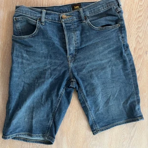 Jeansshorts från Lee - Klassiska blå jeansshorts från Lee med fem fickor och normal passform. Perfekta för sommaren, tillverkade i slitstarkt denimtyg med knapp och dragkedja framtill. Tidlös design med Lee-logga på bakfickan.