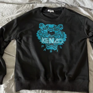 Svart sweatshirt med blått tigertryck från Kenzo - Kenzo sweatshirt i svart med stort blått tigertryck och texten 'KENZO PARIS' framtill. Tröjan har rund halsringning och ribbade muddar vid ärmslut och nederkant. Perfekt för dig som gillar streetwear och vill sticka ut snyggt. Ny pris 1200. Storlek s kommer med boxen och är helt ny har testat den 2 gånger skriv om du är intresserad vi kan prata om priset:)
