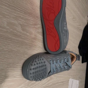 Grå sneakers med nitar från Christian Louboutin - Säljer ett par gråa sneakers från Christian Louboutin med ikoniska röda sulor och nitar på tån. Skorna har snörning och är tillverkade i skinn. Perfekta för dig som vill sticka ut med en edgy och lyxig stil.
