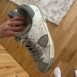 Vita sneakers från Philippe Model - Säljer ett par vita Philippe Model sneakers med grå detaljer och blå hälflik. Skorna har snörning, rund tå och platt sula. Märkets logga syns på sidan och de har en klassisk, sportig look. De har några defekter därav priset! Hör av er om mer🙌