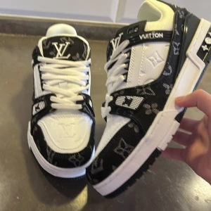 Louis Vuitton sneakers i svart och vitt - Säljer ett par Louis Vuitton sneakers i svart och vitt med tydliga LV-loggor och monogrammönster. Skorna har snörning, rund tå och platt sula. Snygga detaljer på sidan och sulan, samt kontrasterande färger och texturer.