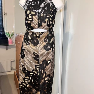 Svart spetsklänning - Elegant svart spetsklänning med vackra blommönster och en öppen rygg. Klänningen har en tight passform och är ärmlös, vilket ger en sofistikerad look. Perfekt för speciella tillfällen.