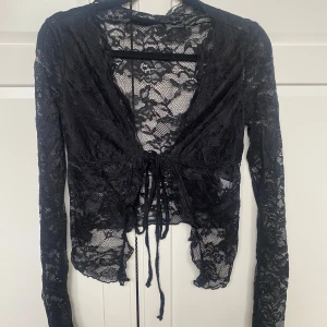 Svart spetsig croppad blus med knyt - Säljer en svart långärmad blus i spets med knyt framtill. Blusen är croppad och har ett transparent blommigt spetsmönster. Perfekt för dig som vill ha en trendig och feminin topp med snygga detaljer.