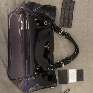 Svart lackad handväska från Prada - Säljer en svart, glansig handväska från Prada i lackat material. Väskan har silverfärgade metalldetaljer, dubbla handtag och medföljande äkthetskort. Klassisk och rymlig modell med exklusiv känsla. Limited edition modell också😝👏 Pris kan diskuteras 👏
