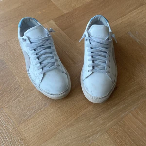 Vita sneakers med ljusblå detalj från Oliver Cabell - Säljer ett par vita sneakers från Common Projects med ljusblå häl och klassisk siluett. Skorna har snörning och är tillverkade i skinn. Perfekta för dig som gillar stilrena och minimalistiska skor.