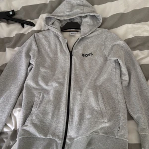 Grå hoodie från BOSS - Snygg grå hoodie från BOSS med dragkedja framtill och huva. Tröjan har två fickor på sidorna och svart BOSS-logga på bröstet. Perfekt för en avslappnad stil och enkel att matcha med andra plagg.