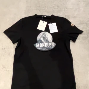 Moncler tshirt storlek L - Säljer en svart t-shirt från Moncler med stort tryck av berg och logga på bröstet samt broderad logotyp på ärmen. Klassisk passform och rund halsringning. Perfekt för dig som gillar stilrena märkesplagg. Den passar en som har storlek L.
