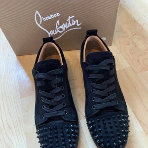 Svarta sneakers med nitar från Christian Louboutin - Säljer ett par svarta sneakers från Christian Louboutin med svarta nitar framtill och klassisk röd sula. Skorna har snörning och är tillverkade i mocka med detaljer i skinn. Perfekta för dig som vill sticka ut med en lyxig och edgy stil. Priset är förhandlingsbart. Allt og ingår