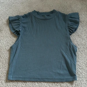 Blå ribbad topp med volangärmar - Supersöt blå ribbad topp med korta volangärmar och rund halsringning. Perfekt att styla till jeans eller kjol för en trendig look.