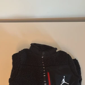 Svart fleecetröja  från Jordan - Säljer en svart fleecetröja  från Jordan med mjukt teddyfoder och svarta paneler framtill. Västen har dragkedja, en ficka med röd dragkedja och klassisk Jordan märke 