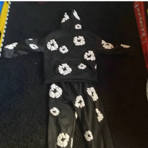 Svart hoodie från Denim Tears med vita blommor - Säljer en svart hoodie från Denim Tears i storlek M. Tröjan har ett unikt vitt blommönster över hela plagget och en klassisk huva. Perfekt för dig som vill sticka ut med en streetwear-look.