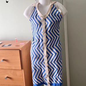 Blå och vit klänning med zigzag-mönster - Säljer en snygg blå och vit klänning med zigzag-mönster. Klänningen har en v-ringad design med knappar framtill och en halterneck-stil. Perfekt för sommarens varma dagar!