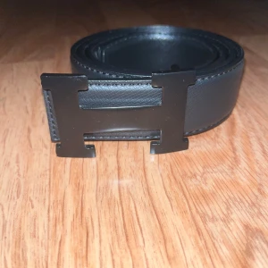 H bälte - Snyggt svart bälte  med ikoniskt H-spänne i metall. Bältet är gjort i äkta skinn med diskret struktur och har stilrena sömmar längs kanterna. Perfekt accessoar för att lyfta din outfit med en lyxig touch. Säljs billigt pga den defekten på den sista bilden. Men ingen som stör om man är försiktigt och går fortfarande lika bra att använda. 110cm