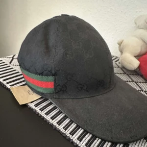 Svart keps från Gucci med GG-mönster - Exklusiv svart keps från Gucci med diskret GG-monogram över hela kepsen och klassiska gröna och röda ränder på sidan. Justerbar baktill och tillverkad i ett slitstarkt material. Snygg och tidlös accessoar som lyfter din outfit.