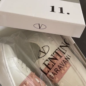 Intressekoll!!Valentino Garavani Rockstud sneakers - Snygga vita Valentino Garavani Rockstud sneakers med rosa band och ikoniska nitar. Supertrendiga och ger en cool edge till din outfit.har använts ganska mycket men ser typ ny ut endå.lite stora för 37 så skulle säga mer 38 i dom.köpte 6000 nya