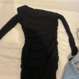 Svart one shoulder långklänning, pris kan diskuteras  - Snygg svart långklänning med one shoulder-design och långa ärmar. Klänningen är tight och har rynkade detaljer längs sidorna för en smickrande passform. Perfekt för dig som vill sticka ut med en elegant och modern look. Ej använd. 