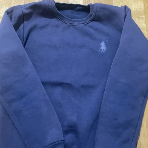Blå sweatshirt från Polo Ralph Lauren - Snygg marinblå sweatshirt från Polo Ralph Lauren med klassisk broderad logga på bröstet. Tröjan har rund halsringning, långa ärmar och ribbade muddar. Perfekt för en chill och stilren look.