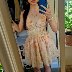 Blommig kortklänning med volangkjol - Supersöt kortklänning med blommigt mönster i ljusa toner av orangea, vit och persika. V-ringning, smala axelband och volangig kjol. Somrigt!🫶