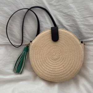 Rund axelväska i strå med tofs - Rund axelväska i beige stråmaterial med svart axelrem. Väskan har en grön tofsdetalj och insidan är fodrad i grönt tyg. Nyskick! 