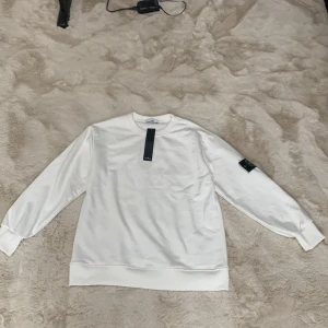 Vit Stone Island sweatshirt - Snygg vit sweatshirt från Stone Island med klassisk rund hals och ribbade muddar. Tröjan har den ikoniska Stone Island-loggan på vänster ärm och är tillverkad i mjuk bomull. Perfekt för dig som gillar stilrena och trendiga plagg.