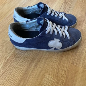 Blå mockasneakers från Gallerist - Snygga blå sneakers i mocka från Gallerist med vita detaljer och snörning. Skorna har en vit klöverdetalj på sidan och vit hälkappa med Gallerist-tryck. Perfekta för dig som gillar en stilren men unik look.