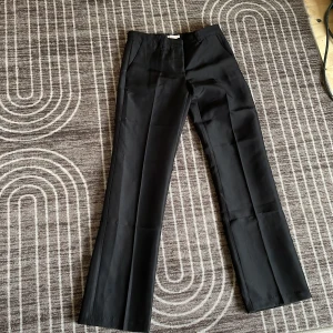 Svarta kostymbyxor från Nelly, stl 34 - Klassiska svarta kostymbyxor från Nelly i storlek 34. Byxorna har raka ben, pressveck och hällor för bälte. Snygg passform med fickor fram och dekorfickor bak. Perfekta för en clean och stilren look. Aldrig använda då dom är för stora för mig 