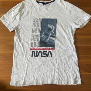 Vit NASA t-shirt med astronauttryck - Vit t-shirt med coolt tryck av en astronaut och NASA-logga på framsidan. T-shirten har rund hals och korta ärmar. Materialet känns mjukt och skönt, perfekt för dig som gillar rymden och vill sticka ut med en unik look.