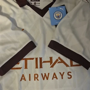 Manchester City vit bortatröja Puma - Snygg Manchester City bortatröja från Puma i vitt med mörkbruna detaljer vid krage och ärmslut. Tröjan har korta ärmar och tryckt Etihad Airways-logga i orange framtill. Perfekt för dig som vill visa vilket lag du håller på.