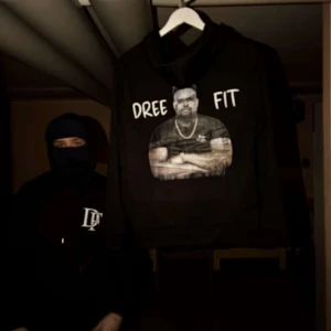Svart hoodie Dree Fit med tryck - Svart hoodie från Dree Fit med stor vit logga på bröstet och ett stort tryck på ryggen med texten 'DREE FIT' och en bild. Klassisk huva med snörning och känguruficka framtill. Perfekt för dig som gillar streetwear och vill sticka ut.