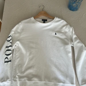Vit Ralph Lauren Tröja - Vit sweatshirt från Polo Ralph Lauren i storlek XL i barn/S Tröjan har rund hals, långa ärmar och svart POLO-tryck längs ena ärmen samt den klassiska lilla broderade loggan på bröstet. Skön och stilren modell i bomull, perfekt för en clean look.