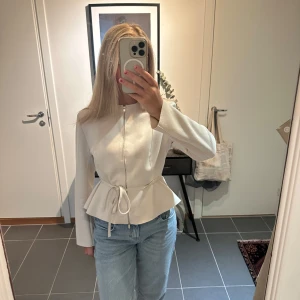 Vit mocha jacka  - Snygg vit kavajjacka med dragkedja framtill och volang nertill. Jackan har ett knytband i midjan som ger en feminin siluett och långa ärmar. Perfekt för dig som gillar stilrena och trendiga plagg.