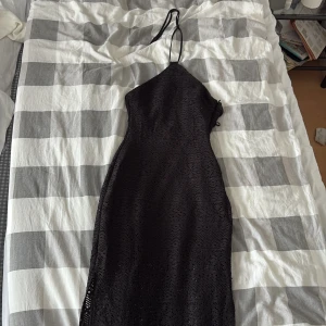 Svart spetsklänning med halterneck - Supersnygg svart långklänning med halterneck och smala axelband. Klänningen har ett vackert spetsmönster och en figurnära passform. Dragkedja i sidan för enkel påklädning. Perfekt för dig som vill sticka ut med en elegant look.