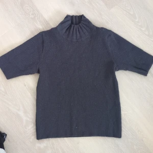 Mörkgrå ribbad polotröja - Ribbstickad polotröja i mörkgrått med hög, bred krage och halvkort ärm. Tröjan har en clean och stilren look, perfekt för lager på lager. Materialet känns mjukt och ger en snygg struktur. Passar dig som gillar minimalistisk och modern stil.