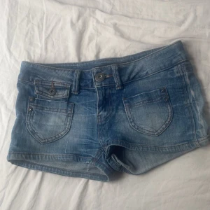Blå jeansshorts från ONLY W27 - Snygga blå jeansshorts från ONLY med klassisk femficksdesign och slitna detaljer. Shortsen har låg midja, knapp- och dragkedjestängning samt coola nitar på fickorna. Perfekta för sommardagar och festivalhäng.