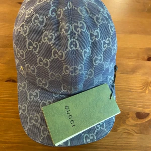 Blå keps från Gucci - Snygg blå keps från Gucci med ikoniskt GG-mönster över hela kepsen. Justerbar rem baktill i blått läder med guldfärgad detalj. Perfekt för dig som vill sticka ut med en lyxig accessoar. Kepsen är icke andvänd alltså helt ny!