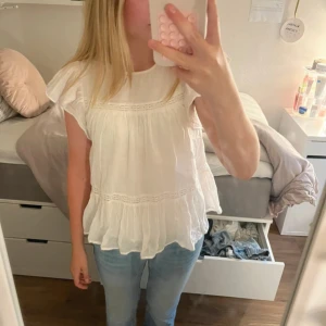 Vit volangblus med spetsdetaljer - Supersöt vit blus med volangärmar och luftigt fall. Blusen har fina spetsdetaljer och en lätt rynkad struktur som ger en romantisk vibe. Perfekt till jeans eller kjol för en fräsch look. Ärmlös modell med rund halsringning.