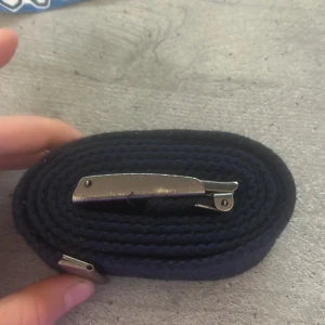 Svart tygbälte med metallspänne - Snyggt svart tygbälte med klassiskt silverfärgat metallspänne. Bältet är gjort i slitstarkt tygmaterial och har en enkel, rak design utan mönster. Perfekt för att ge en clean look till jeans eller chinos.
