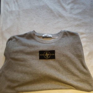 Grå Stone Island sweatshirt - Snygg grå sweatshirt från Stone Island med klassisk rund hals och ikonisk svart patch med gult broderat kompassmärke på bröstet. Tröjan är i mjuk bomull och har en clean, stilren look som passar perfekt till jeans eller trackpants.