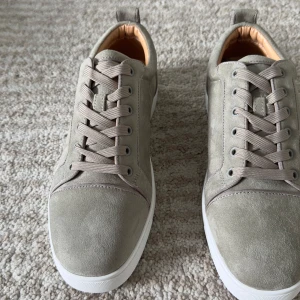 Grå mockasneakers från Christian Louboutin - Snygga sneakers i beige mocka från Christian Louboutin med klassisk röd sula. Skorna har rund tå, platt vit sula och ton-i-ton snörning. Insidan är i mjukt skinn för extra komfort. Perfekta för dig som vill ha en clean och exklusiv look.