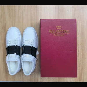 Valentino Garavani Open sneakers vit/svart - Snygga Valentino Garavani Open sneakers i vitt skinn med bred svart rem över vristen. Klassisk rund tå, platt sula med nitar baktill och diskret logga på plösen. Perfekta för dig som vill ha en clean men edgy look.