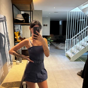 Mörkblå kort klänning med bälte - Säljer en mörkblå kort klänning med smala axelband och markerad midja med matchande bälte. Klänningen har en figurnära passform upptill och lätt utställd kjol med shorts under. Perfekt för dig som gillar en stilren och trendig look.