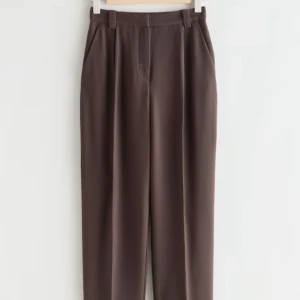 Bruna kostymbyxor & Other Stories - Snygga bruna kostymbyxor från & Other Stories med hög midja och pressveck. Byxorna har raka ben, uppvikta benslut och hällor för bälte. Perfekta för en clean och stilren look.