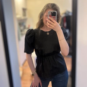 Svart blus med puffärmar - Snygg svart blus med puffiga korta ärmar och markerad midja. Blusen har rund halsringning och ett lätt glansigt tyg som ger en elegant känsla. Perfekt att styla med jeans eller kjol för en trendig look.