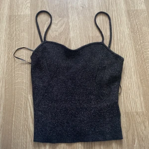 Svart glittrig linne-topp - Supersnygg svart linne-topp med tunna axelband och glittrigt tyg. Toppen har en lätt figurnära passform och en söt, svagt hjärtformad urringning.  Har bara haft på mig två gånger så den är i jätte bra skick.💕