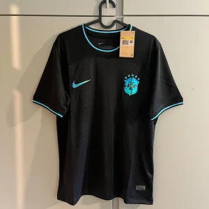 Svart Brasilien fotbollströja Nike - Snygg svart Brasilien fotbollströja från Nike med blå detaljer och CBF-emblem på bröstet. Tröjan har korta ärmar, rund hals och diskret mönster i tyget. Tillverkad i lätt och ventilerande Dri-FIT-material som passar perfekt för träning eller match.